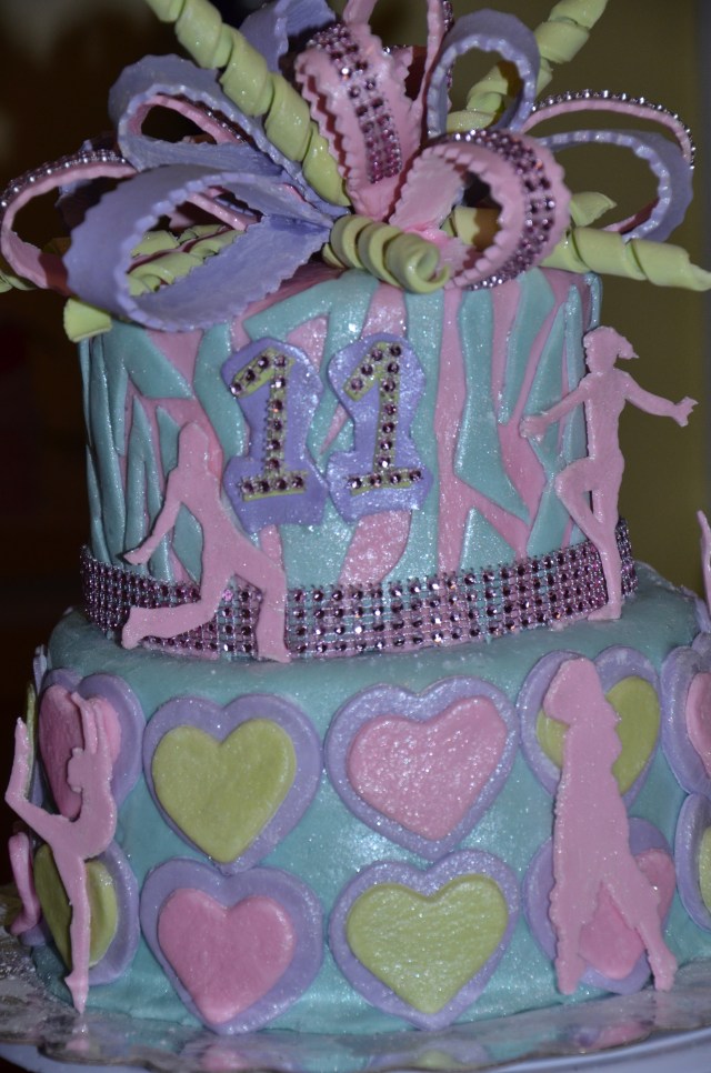  A By Grace Cakes birthday cake original. Gluten-free chocolate cake with vanilla buttercream. Covered with extra sparkles this cake displays hip hop and gymnastics silhouettes and a multiloop, multi-color bow.